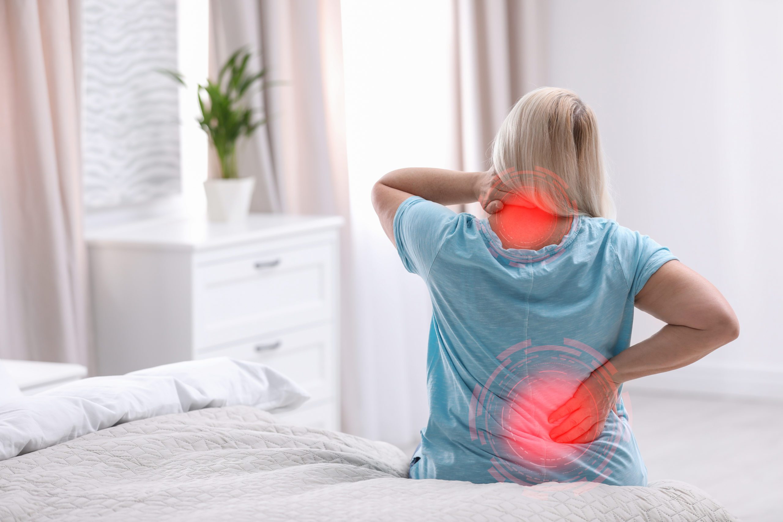 A woman sitting on the edge of a bed holding her neck and lower back, highlighted areas suggesting chronic pain management, in a brightly lit bedroom.
